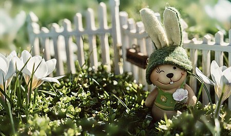 Eine Osterhasenfigur steht vor einem Miniaturzaun zwischen Krokusblüten