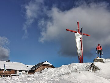 Neben einer Hütte steht ein angeschneites Gipfelkreuz und daneben steht eine Wanderin mit Schneeschuhen