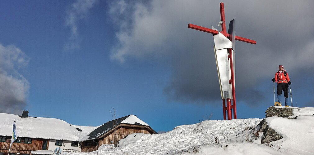Neben einer Hütte steht ein angeschneites Gipfelkreuz und daneben steht eine Wanderin mit Schneeschuhen